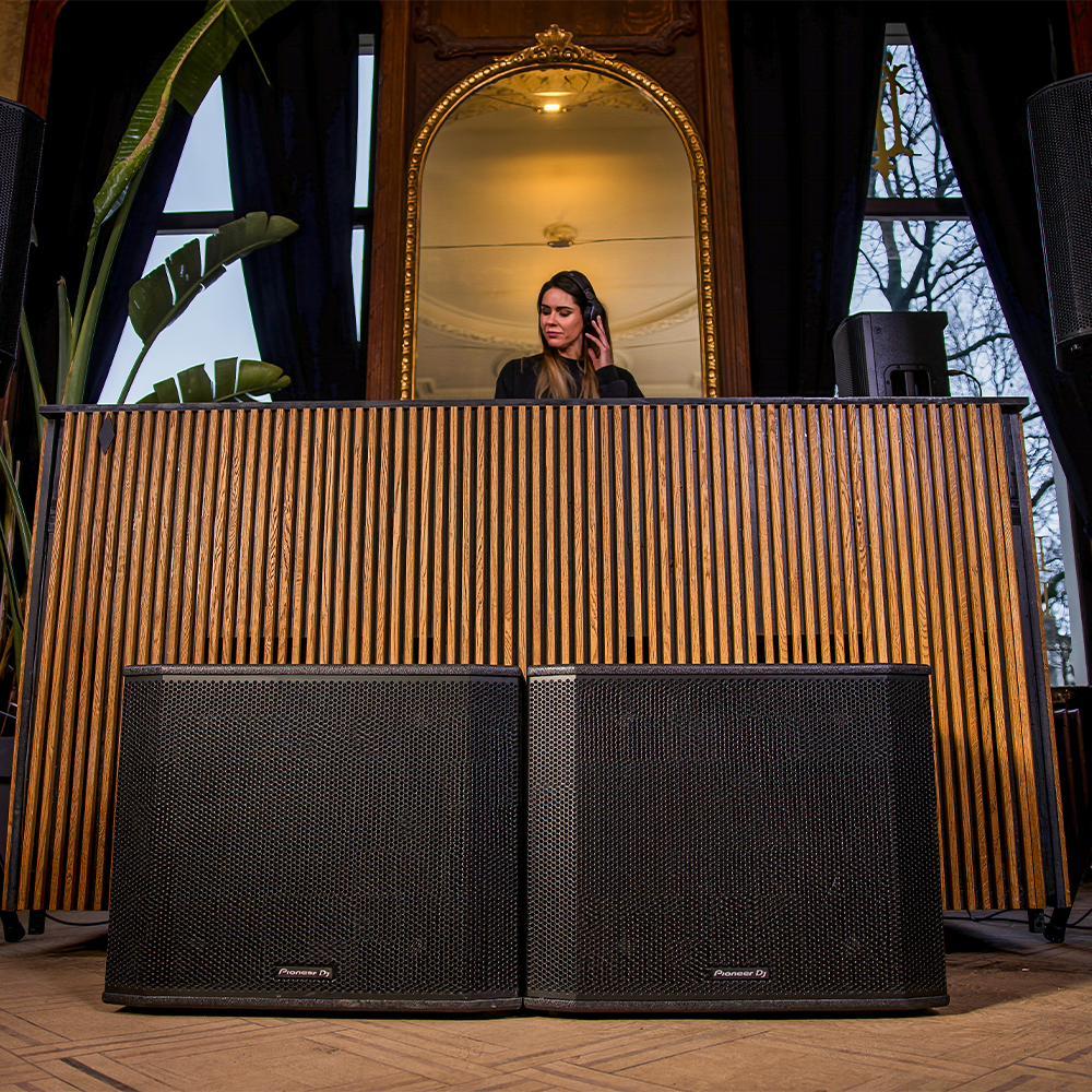 Dos subwoofers Pioneer DJ XPRS2 colocados bajo una cabina de DJ en una sala