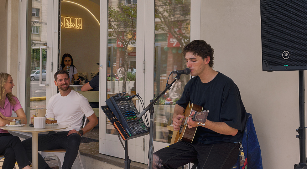 Cantautor con guitarra y mesa de mezclas en la terraza de una cafetería, con un Mackie Thump12v4 sobre trípode